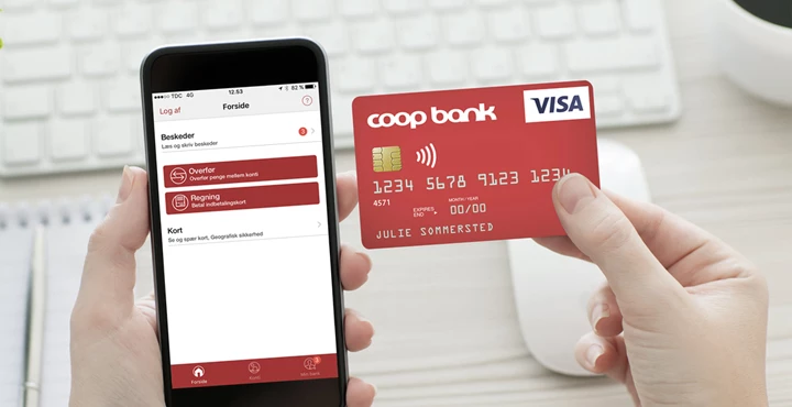 En person holder en smartphone med Coop Bank-app og et rødt Coop Bank Visa-kort i hænderne.