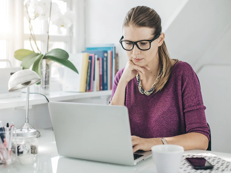 Kvinde i lilla trøje og briller arbejder koncentreret på laptop ved skrivebord.