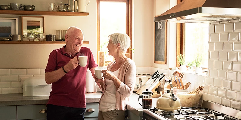 Ældre par i køkkenet, der smiler og drikker kaffe sammen ved siden af køkkenapparater.
