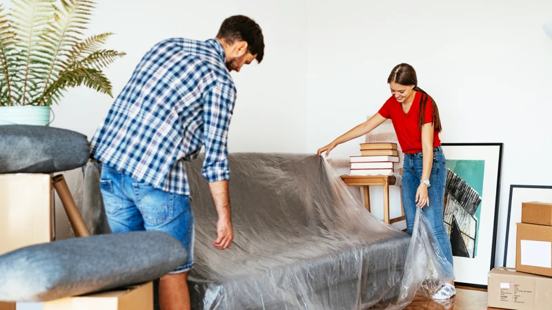 Mand og kvinde dækker en sofa med plastfolie i et rum med flyttekasser og kunstværker.