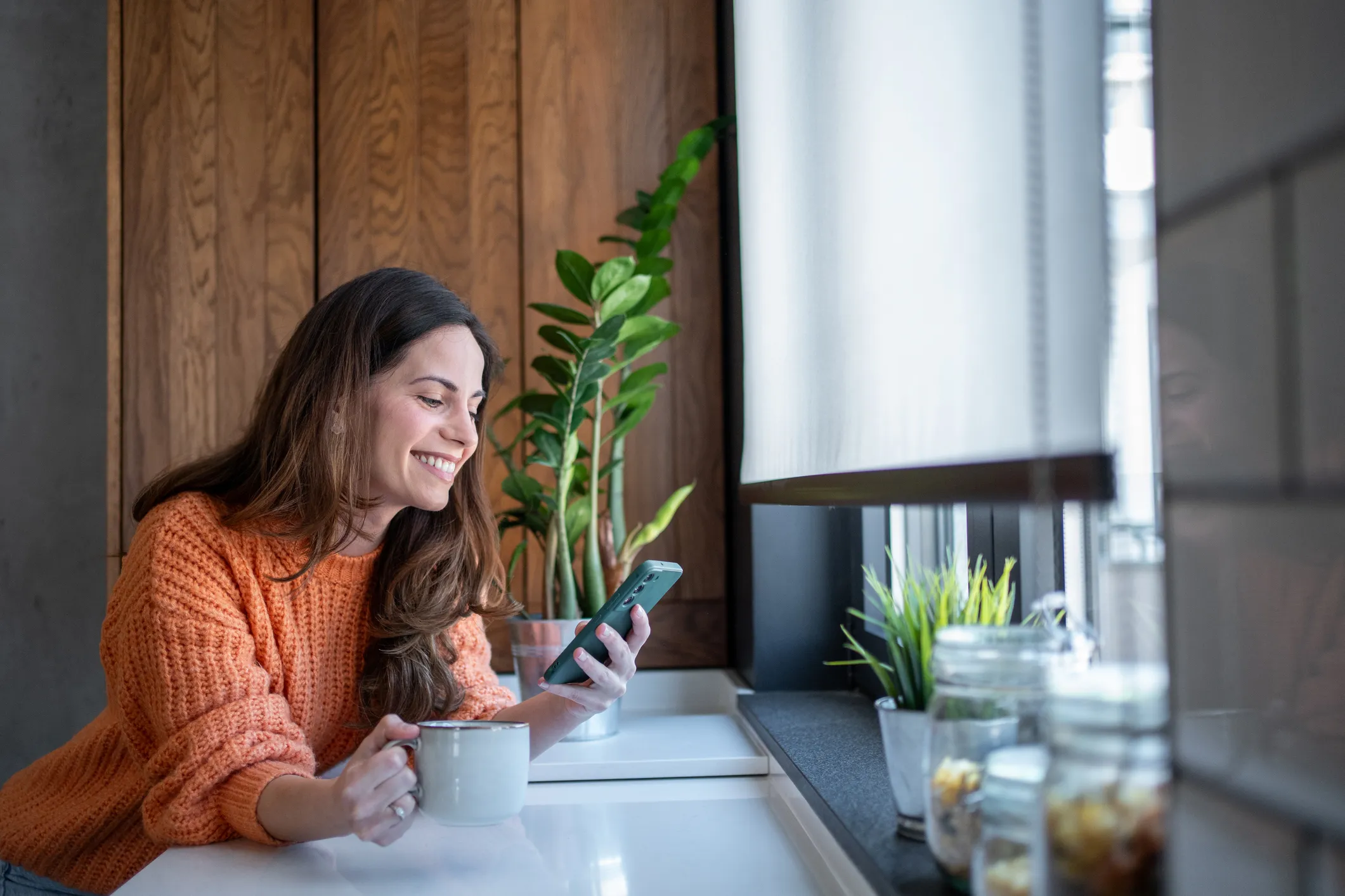 Glad kvinde i orange sweater, der holder en kop, mens hun ser på sin telefon ved vinduet.