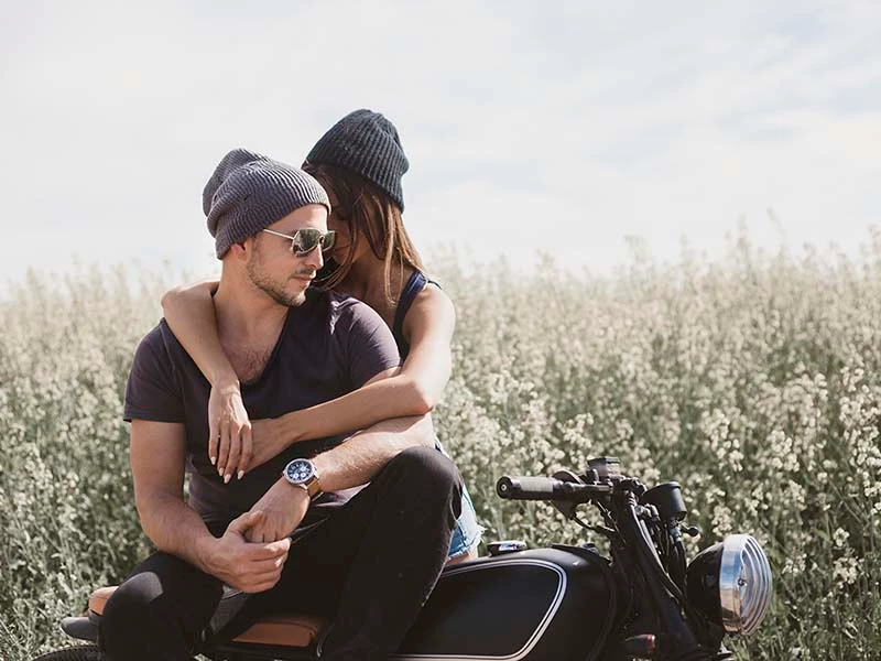 Par kærester i strikhatte krammer på en motorcykel i en blomstrende mark.