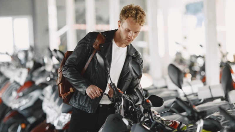 Mand med sort læderjakke og rygsæk tjekker motorcykel i butik med flere motorcykler.