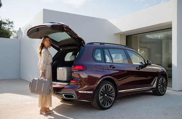 Kvinde med kufferter lægger bagage i bagagerummet på en mørkerød SUV parkeret ved et moderne hus.
