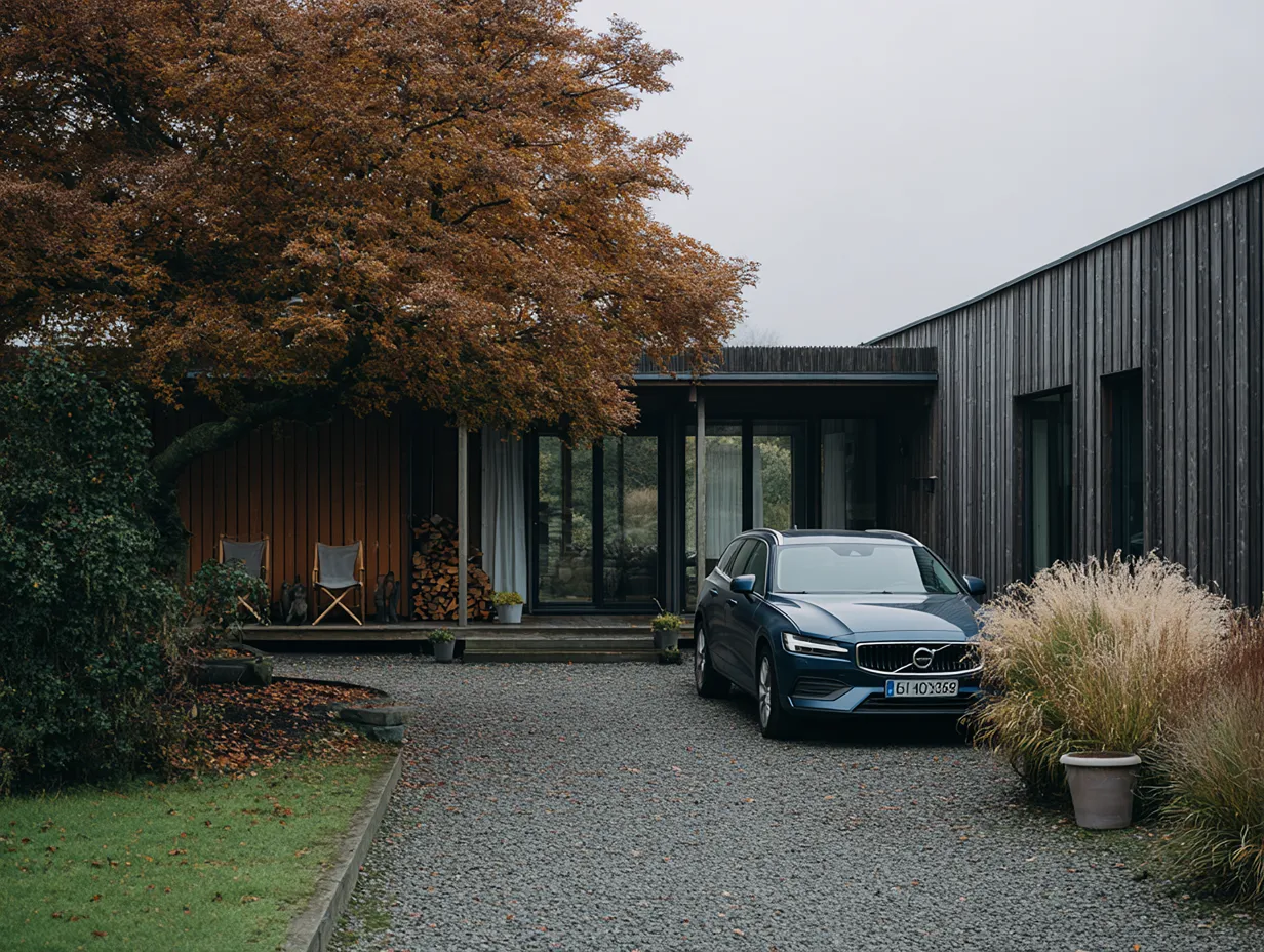 Blå Volvo parkeret foran moderne træhus med store efterårsfarvede træer og græsplæne.