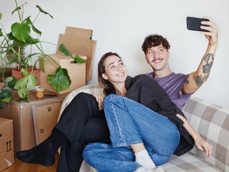 Et par smilende sidder sammen på en sofa og tager en selfie med en mobiltelefon.