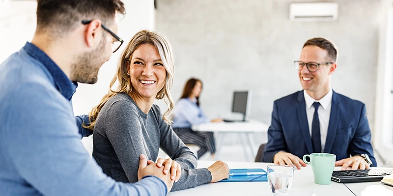 Tre professionelle personer smilende og samtalende ved et kontorbord med computere og kopper.