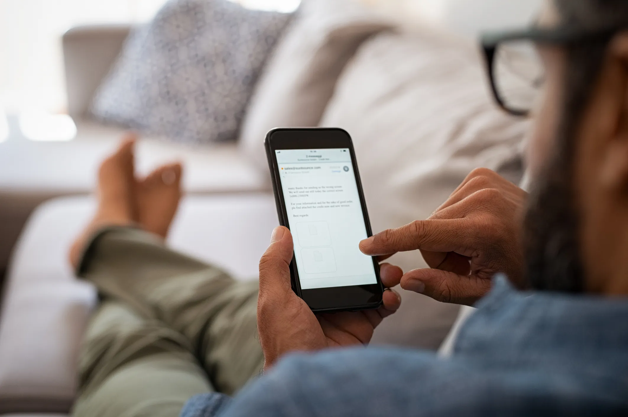 Person med briller sidder på en sofa og læser en e-mail på en smartphone.