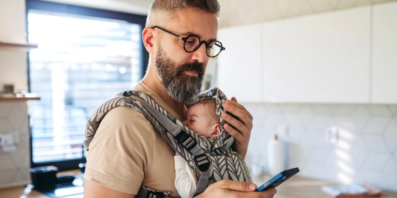 Mand med skæg og briller bærer sovende baby i bæresele og ser på smartphone i køkken.