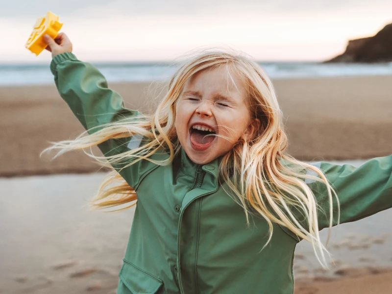 Glad lille pige med langt blondt hår bærer grøn jakke og holder gul klods på stranden.