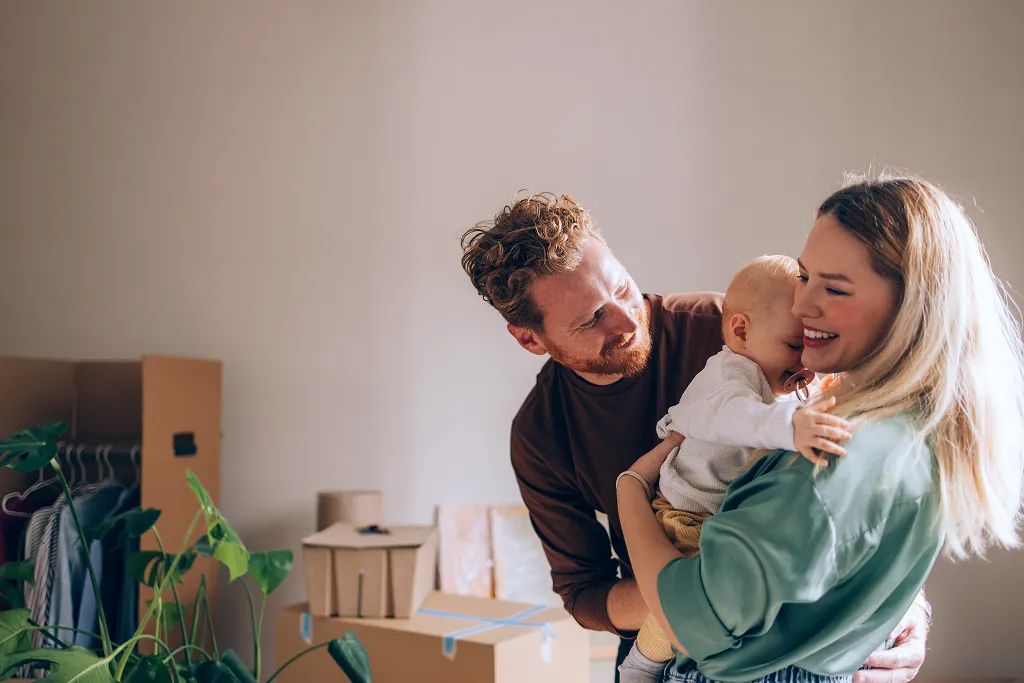 Glade forældre holder baby og smiler i et rum med papkasser og planter.