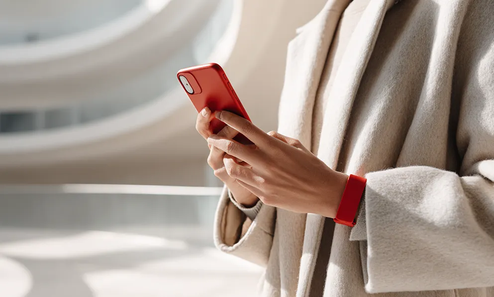 Person iført beige frakke bruger mobiltelefon med rødt cover og matchende rødt armbånd.