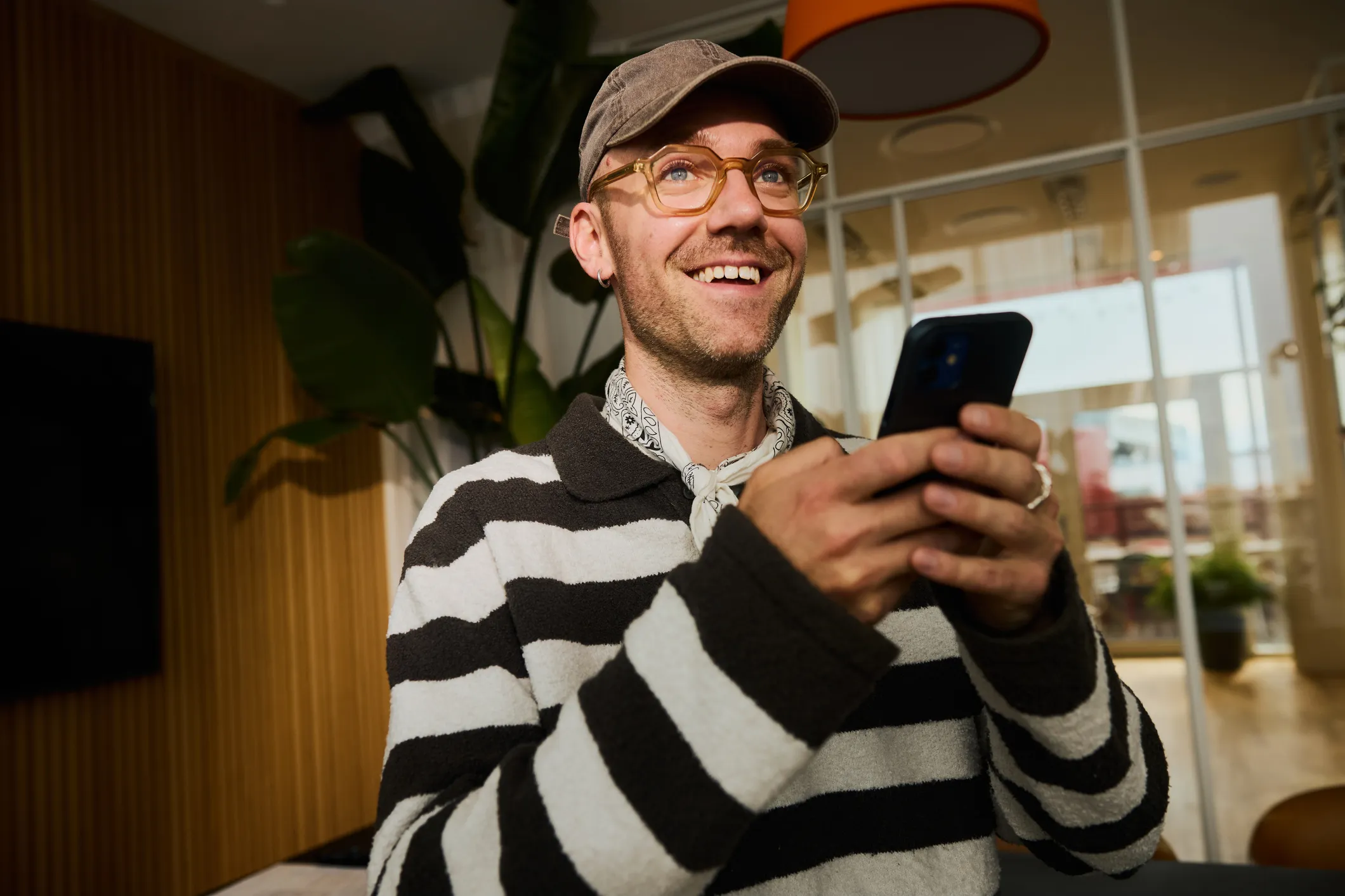 Smilende mand med stribet sweater, kasket og briller, der holder en smartphone indendørs.