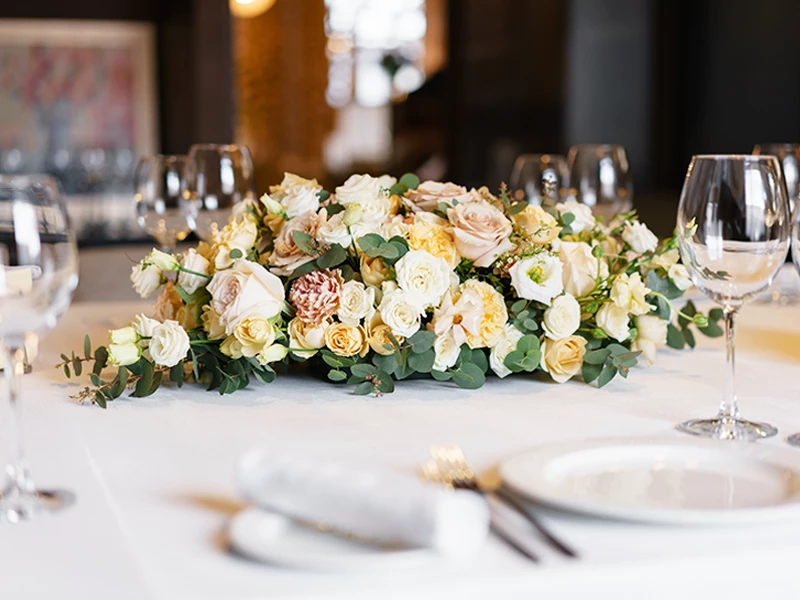 Elegant blomsterarrangement med hvide og rosa roser på et dækket bord med glas og porcelæn.