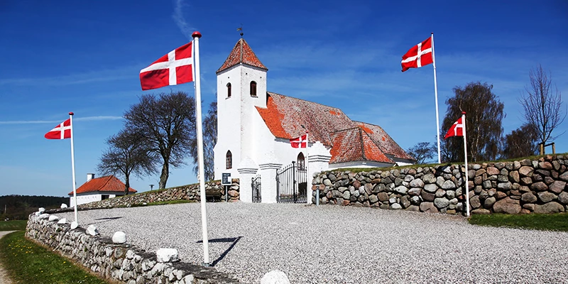 Hvid kirke med rødt tag omkranset af fire danske flag ved en stenet indkørsel.