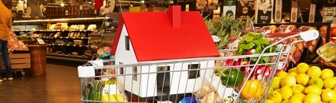 Indkøbsvogn med en model af et hus med rød tag blandt frugt og grøntsager i butik