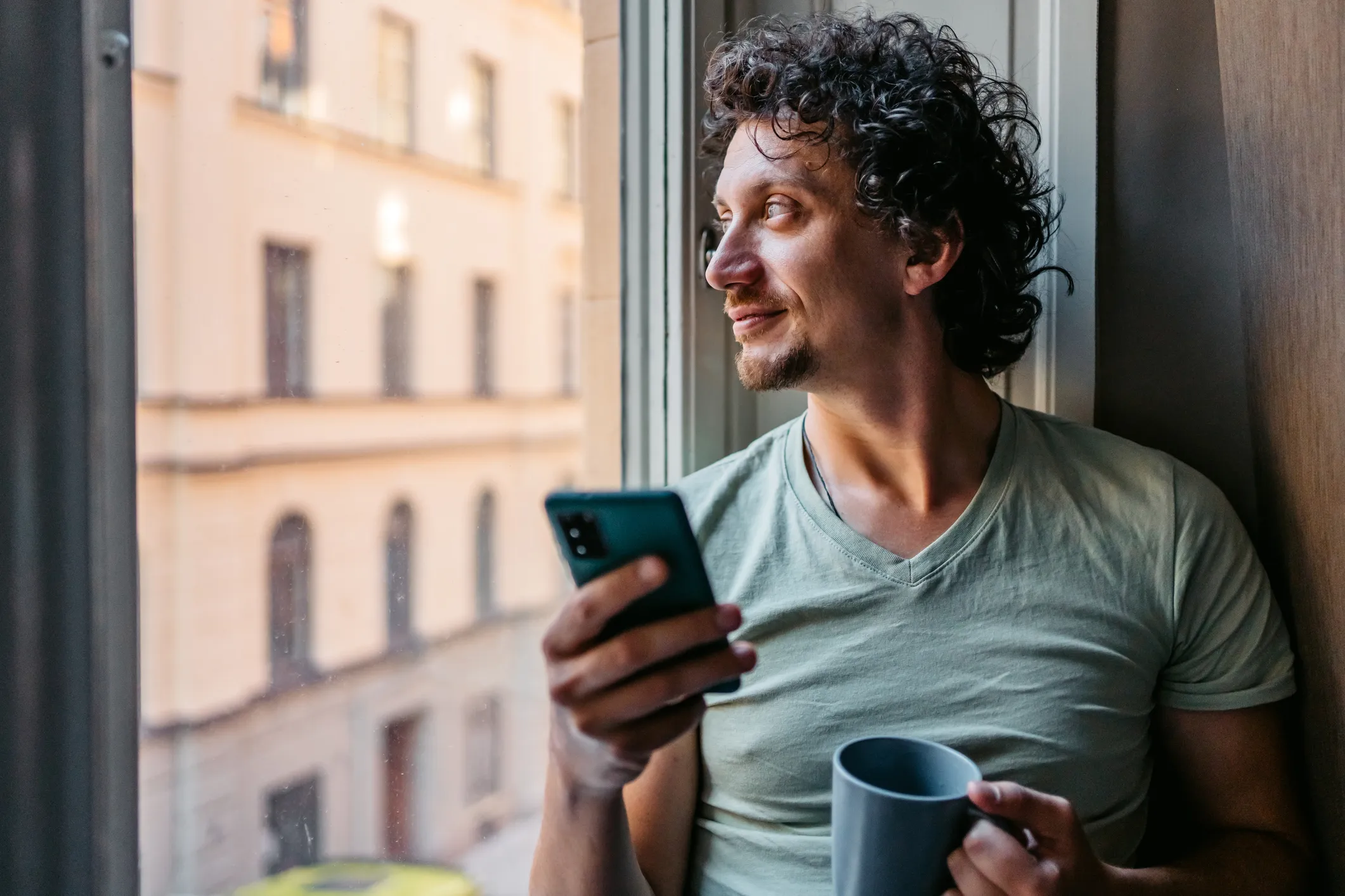 Mand med krøllet hår ser ud af vindue med telefon i den ene hånd og kop i den anden.