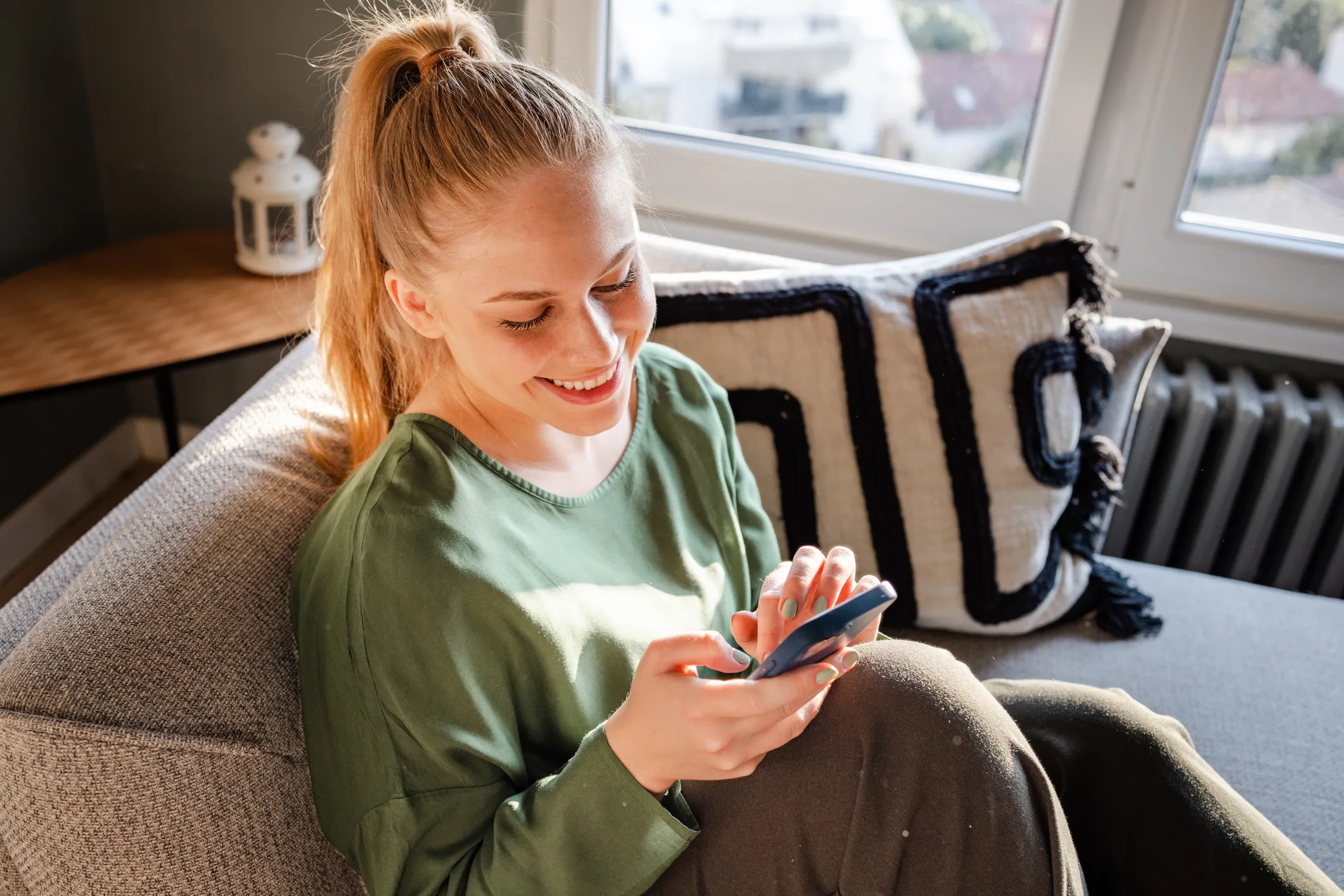 Glad ung kvinde sidder på sofa og smiler, mens hun bruger en smartphone.