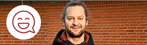 Mand med knold og sort jakke smiler foran en murstensvæg med en smilende emoji ved siden af.