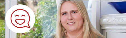 Smilende blond kvinde sidder foran vindue med grønne træer udenfor.