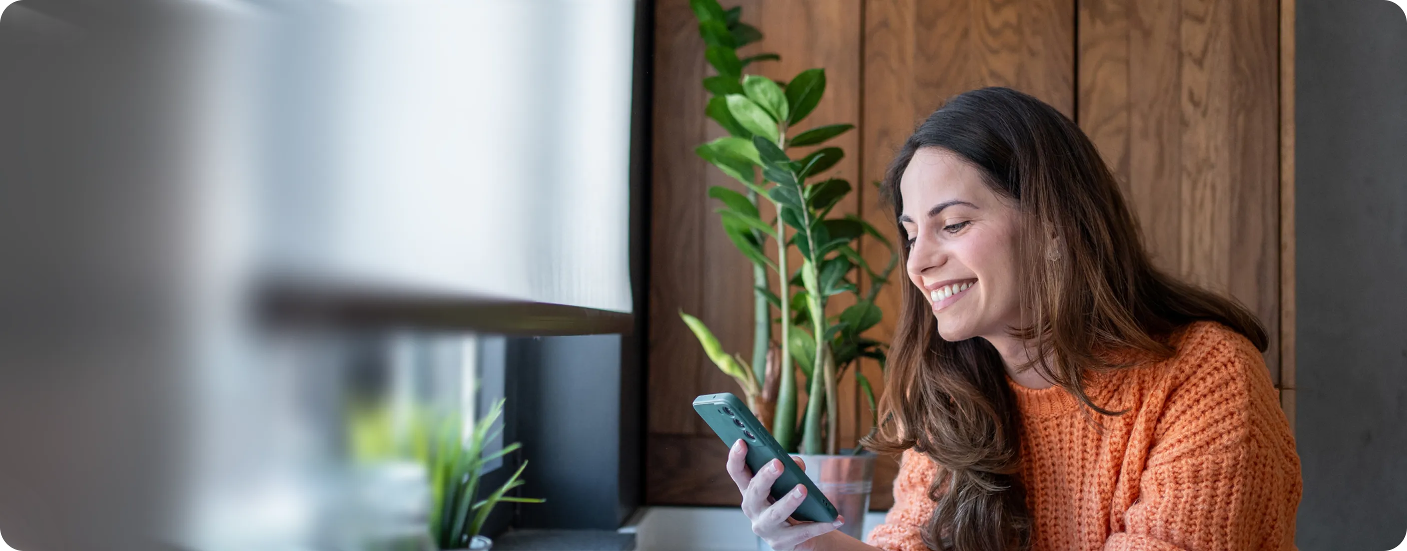 Smilende kvinde i orange sweater ser på sin smartphone indendørs ved vindue med planter.
