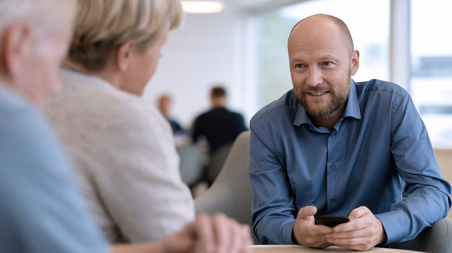 Mand i blå skjorte taler venligt med to ældre personer ved et bord i et lyst rum.