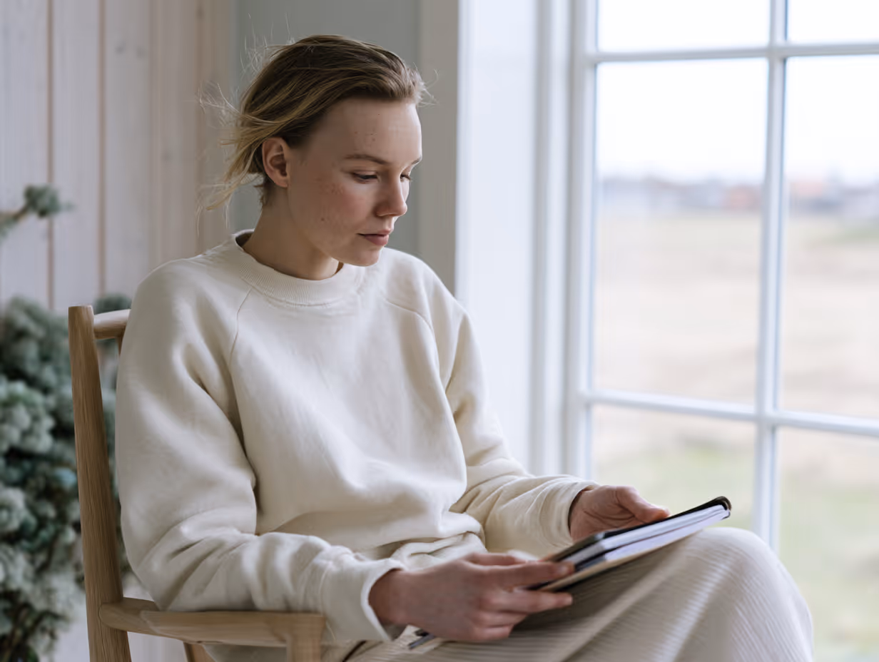 Kvinde iført en hvid sweater, der sidder i en træstol ved et vindue og læser på en tablet.