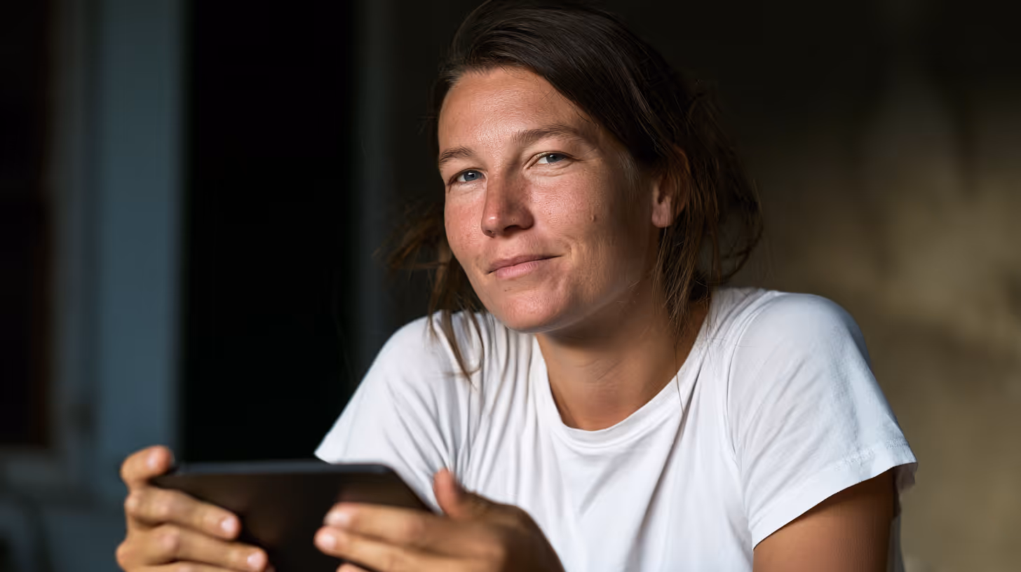 Kvinde med hvid t-shirt, der holder en mobiltelefon og kigger ind i kameraet med et let smil.