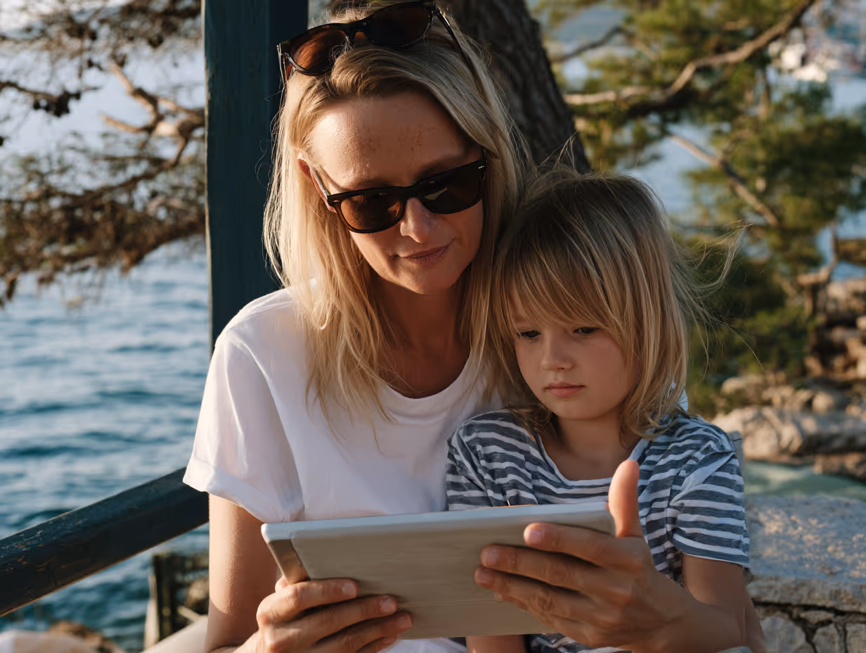 Kvinde med solbriller og barn kigger koncentreret på en tablet udendørs ved vandet.