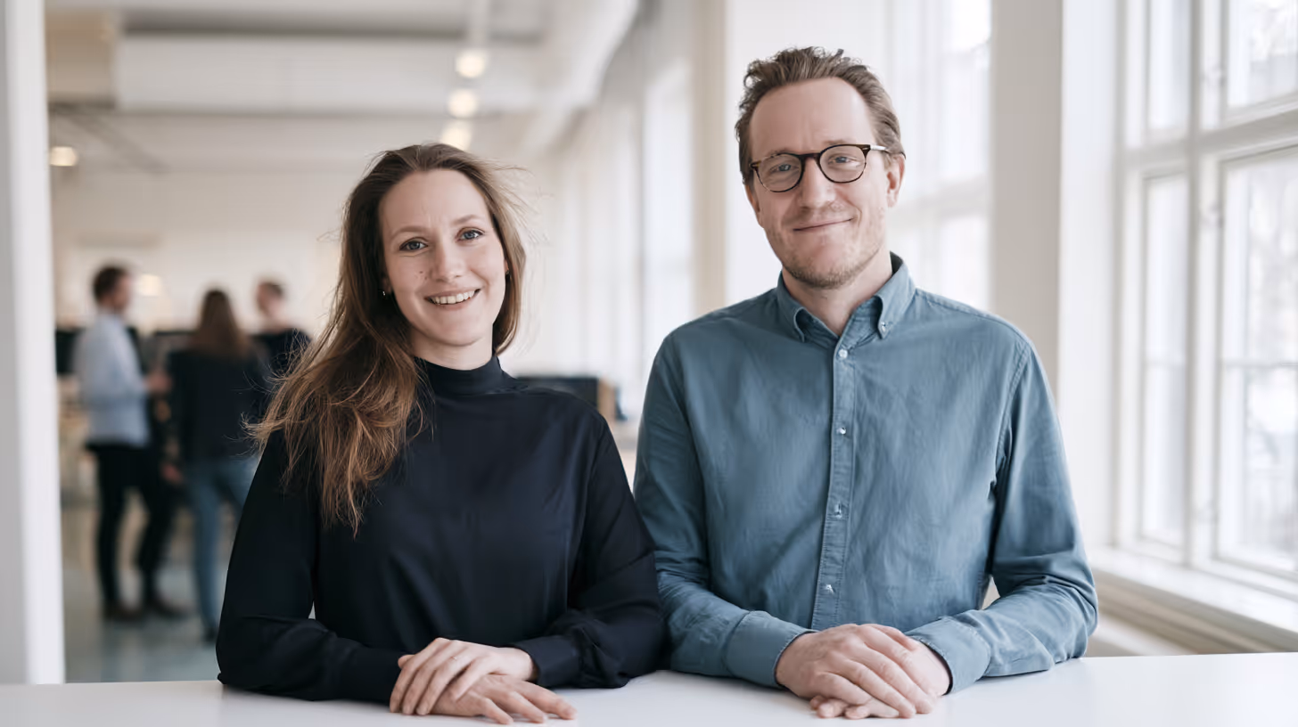 To smilende personer sidder ved et hvidt bord i et lyst, moderne kontor med folk i baggrunden.