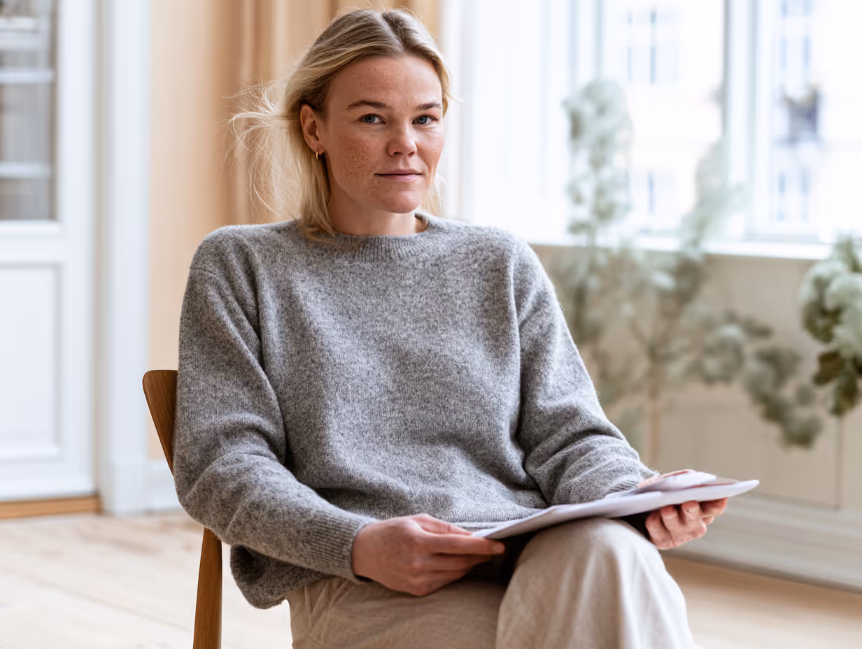 Kvinde i grå sweater sidder på stol med papirer i et lyst rum med store vinduer.