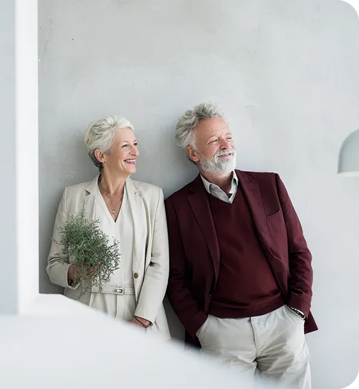 Modent par smilende, hun holder en grøn plante, begge i elegante jakkesæt mod hvid væg.
