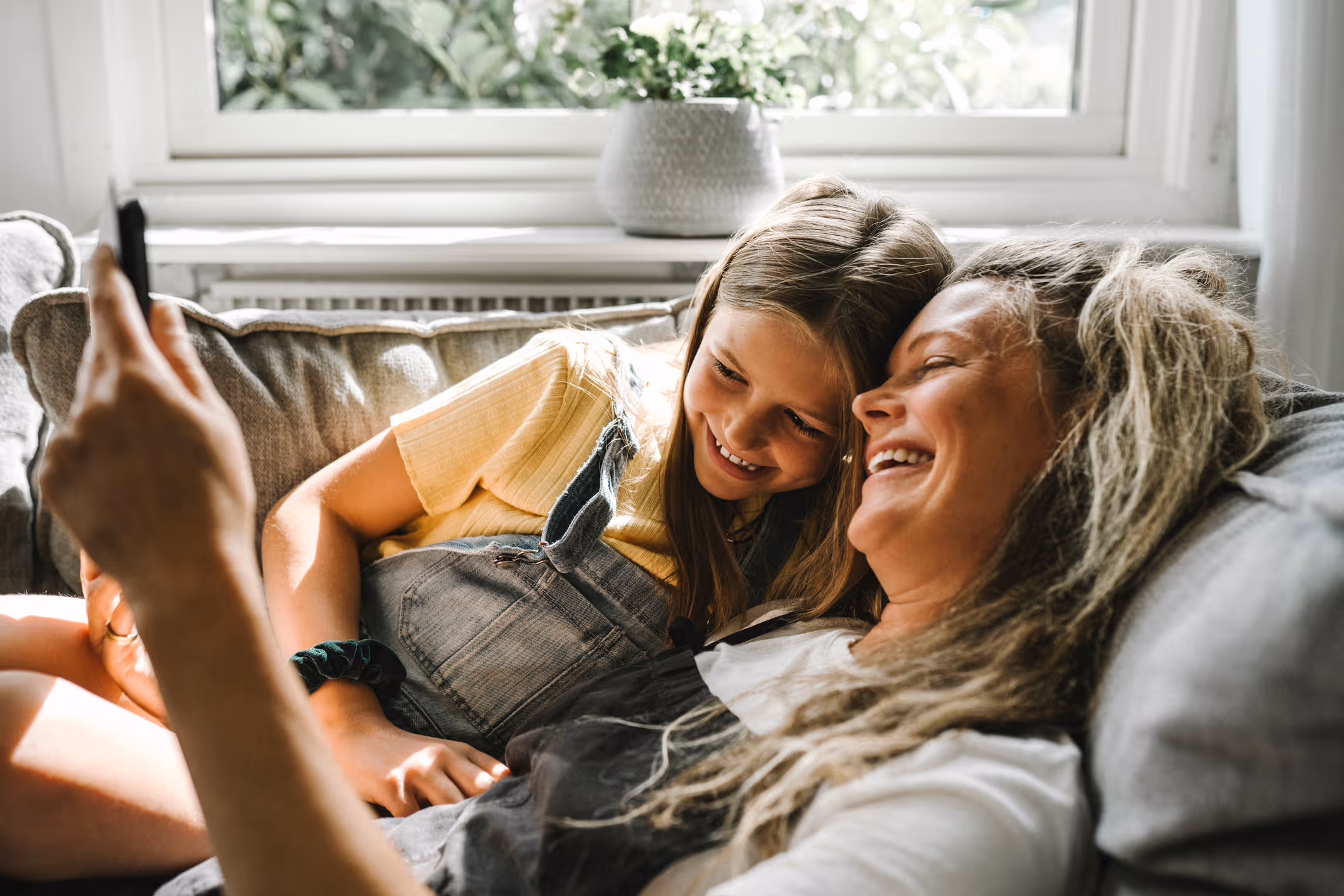 Mor og datter ligger sammen på en sofa og smiler, mens de tager en selfie med en smartphone.