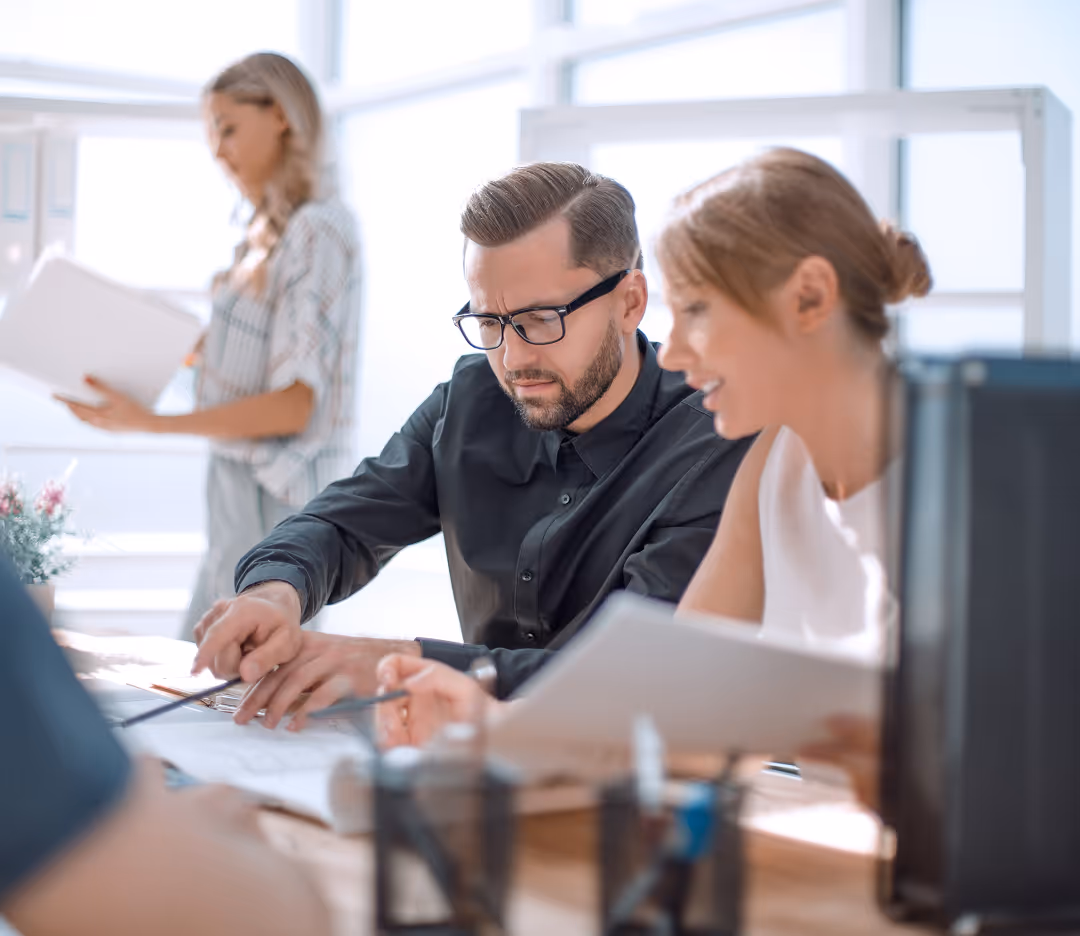 Two colleagues reviewing documents together at a desk while a woman stands reading papers in the background.