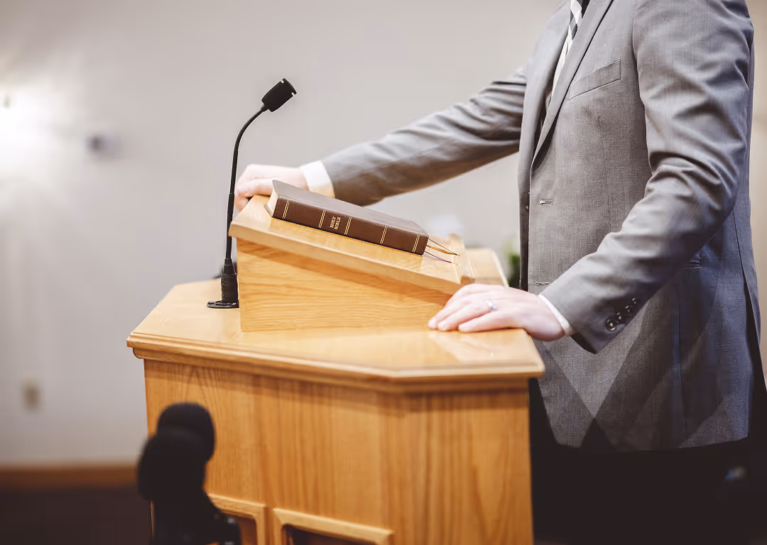 Person in gray suit standing at a wooden podium with a microphone and a closed Bible.