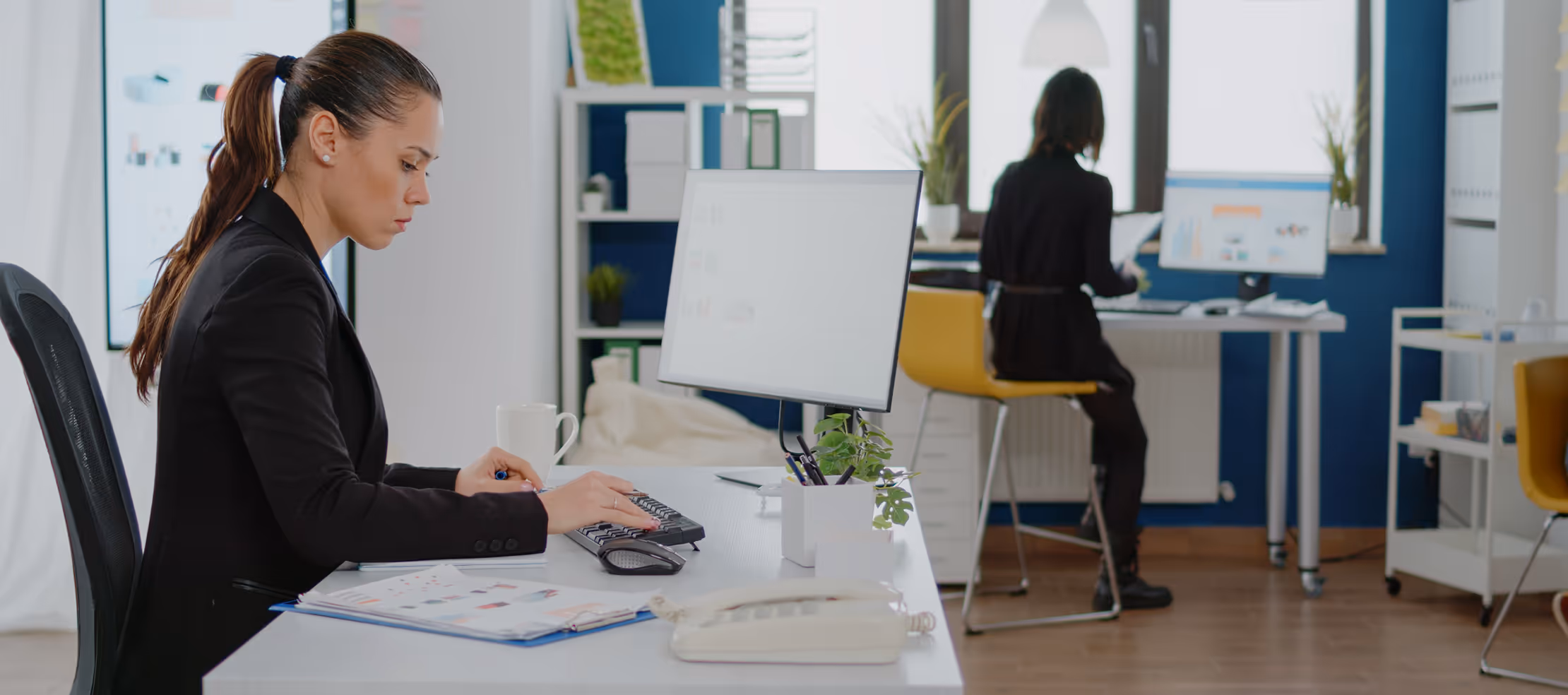 Two women working in an office; one types on a keyboard at a desk in the foreground, the other reads documents near a window in the background.