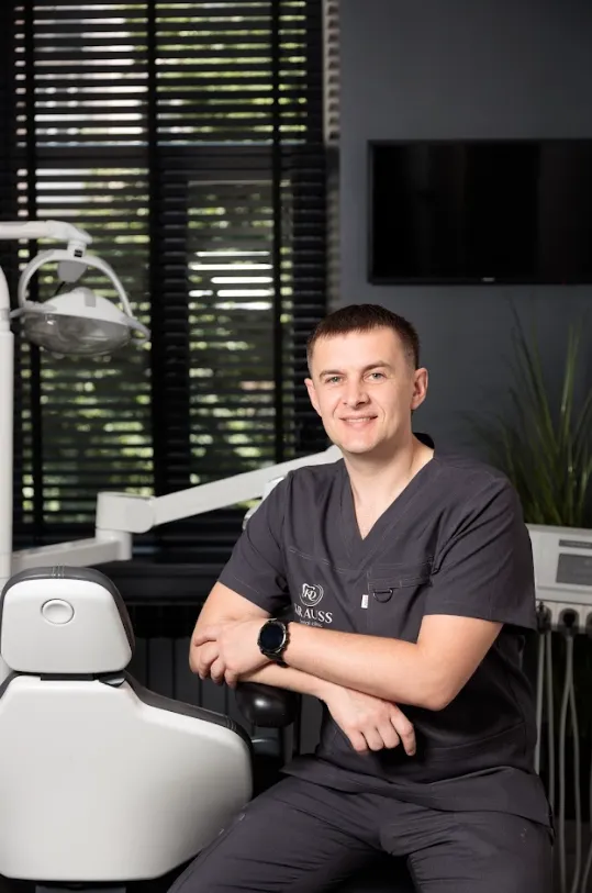 A male dentist in dark scrubs sitting and smiling beside a dental chair in a modern dental office.