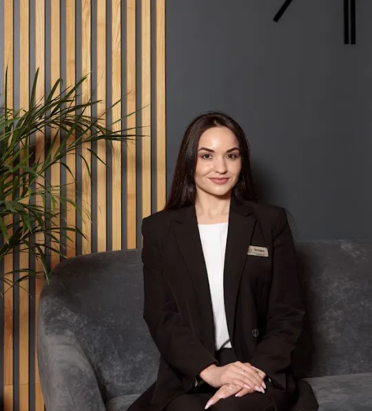 Smiling woman with long dark hair in a black blazer sitting on a gray sofa with a wooden panel and plant in the background.