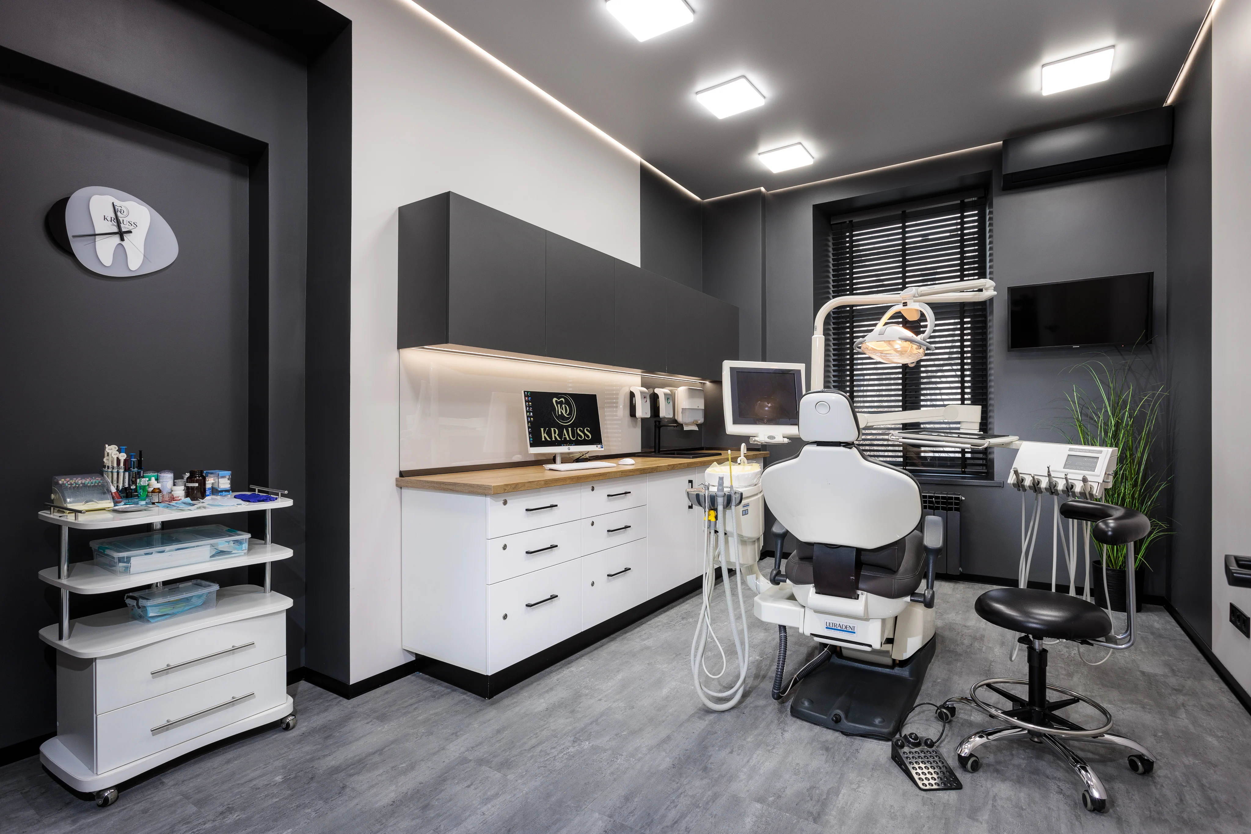 Modern dental clinic room with a dental chair, equipment, computer, and a rolling cart with medical supplies.