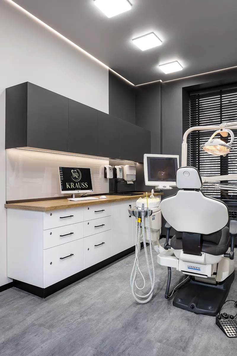 Modern dental clinic room with dental chair, overhead light, equipment, and a computer displaying the Krauss logo.