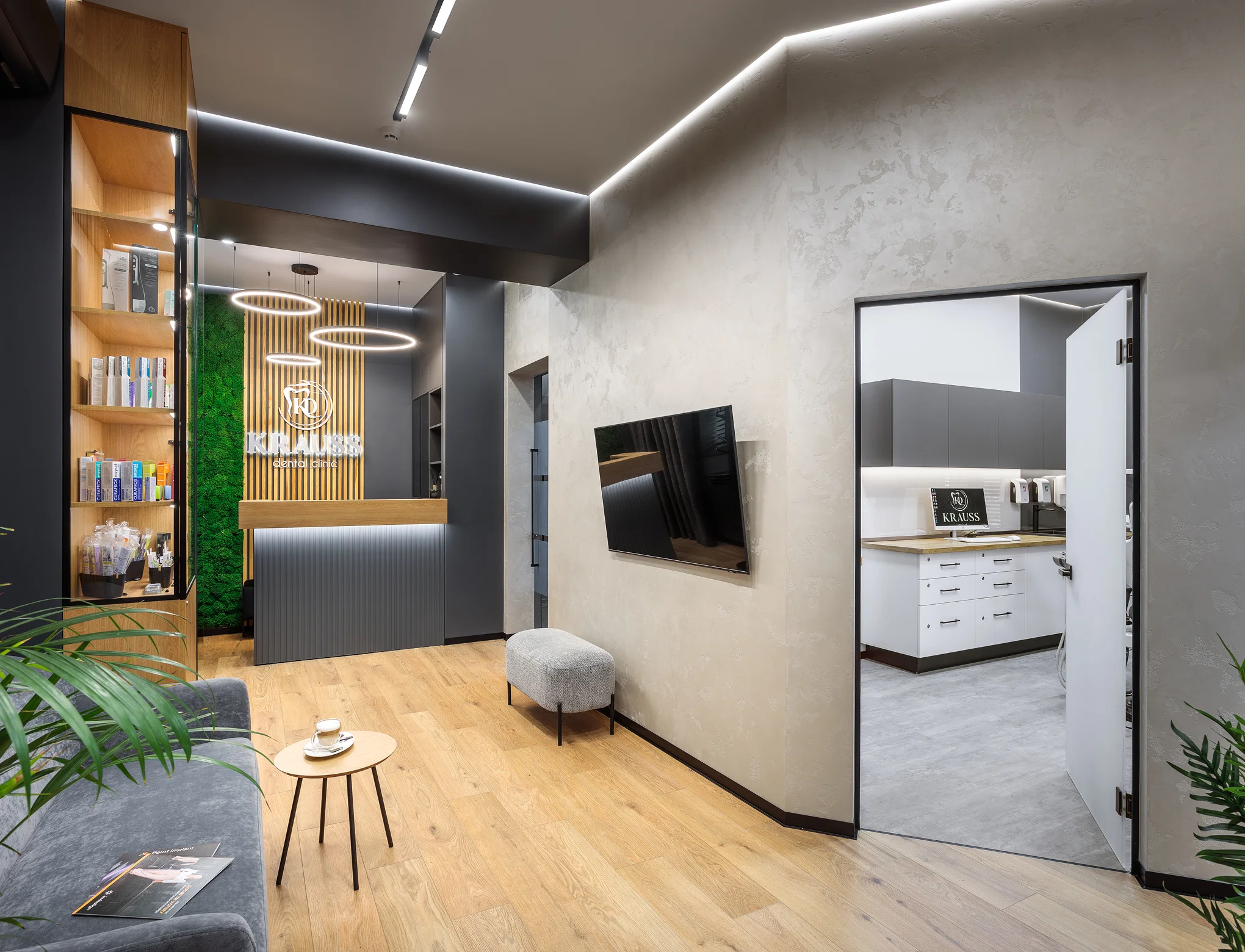 Modern dental clinic reception area with a grey couch, wooden floor, ring pendant lights, and an open door leading to a clinical room.
