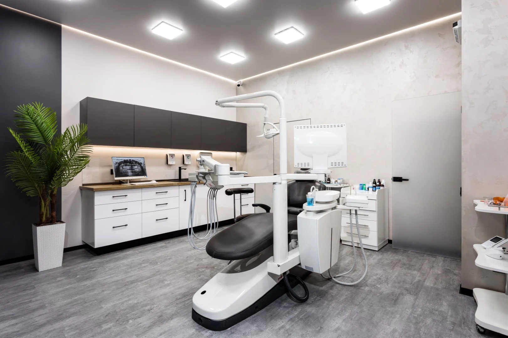 Modern dental office with black and white dental chair, cabinetry, computer screen showing dental X-ray, and decorative plant.