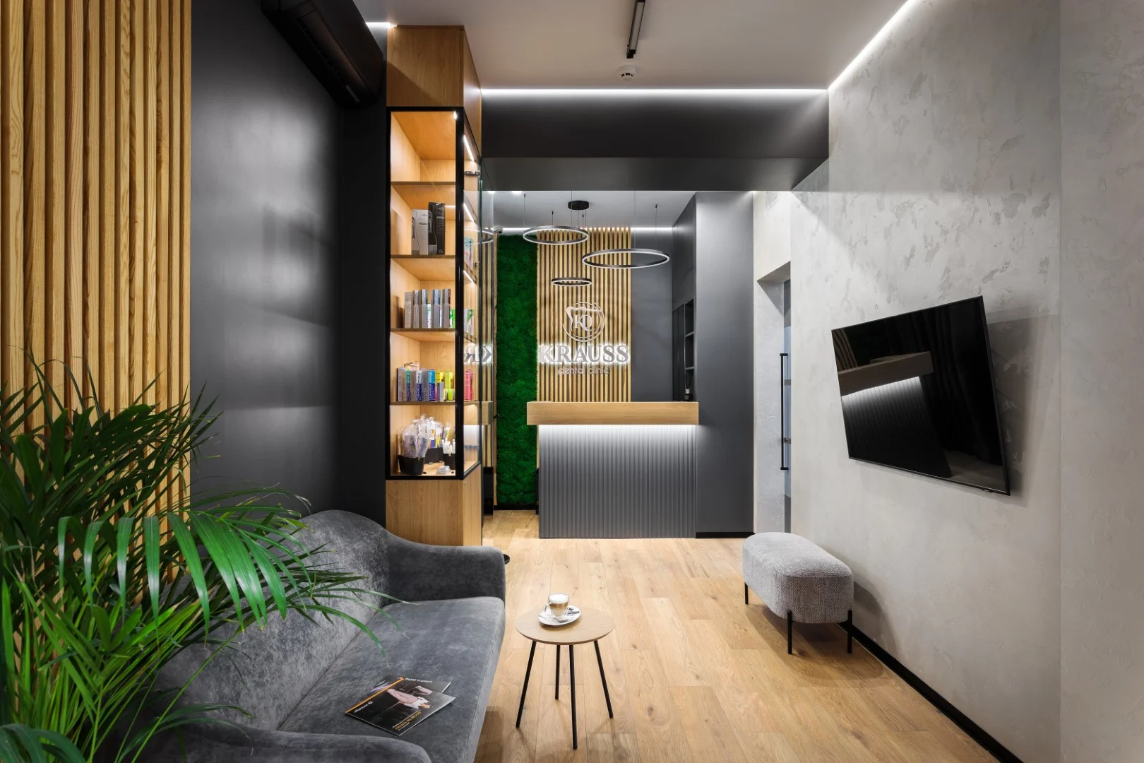 Modern dental clinic waiting room with grey sofa, small wooden table with coffee, TV mounted on wall, and reception desk with illuminated KRAUSS sign.