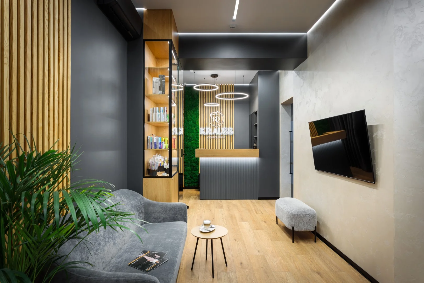 Modern dental clinic waiting area with gray sofa, small round wooden table, wall-mounted flat-screen TV, and reception desk with illuminated logo.