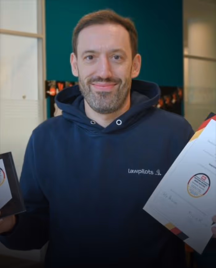 Steffen Hetzel wearing a navy hoodie with 'lawpilots' logo holding two certificates and smiling at the camera.