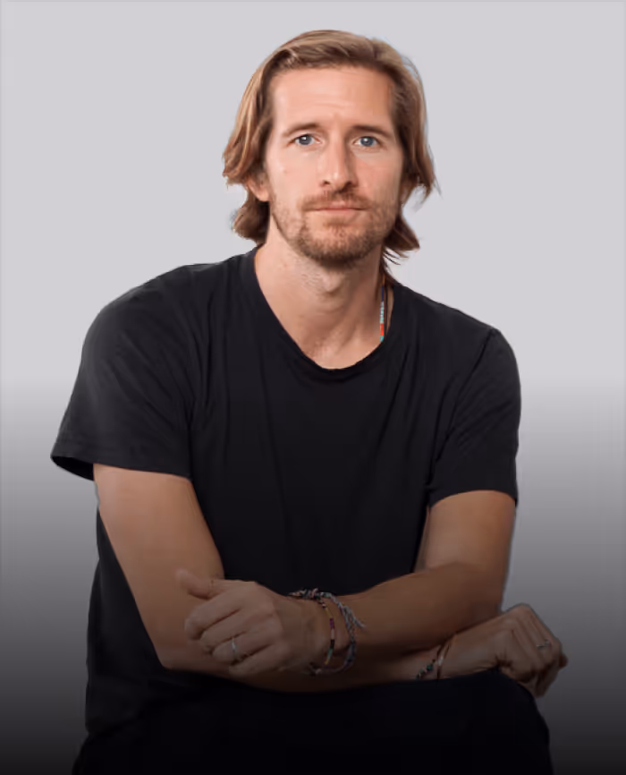 Jakob Schreyer with medium-length light brown hair and a beard, wearing a black t-shirt, sitting with arms crossed.