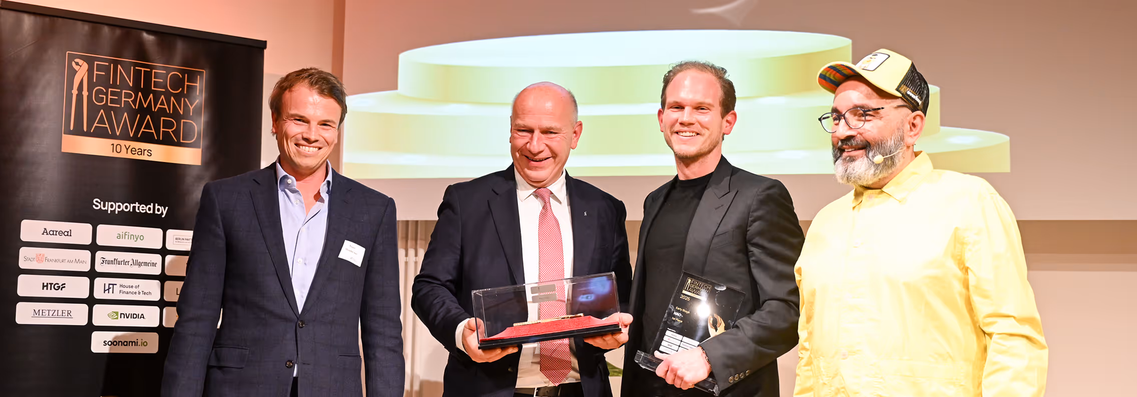 Vier lächelnde Männer auf einer Preisverleihung, zwei halten Auszeichnungen, links steht ein Banner mit Fintech Germany Award 10 Years Logo.