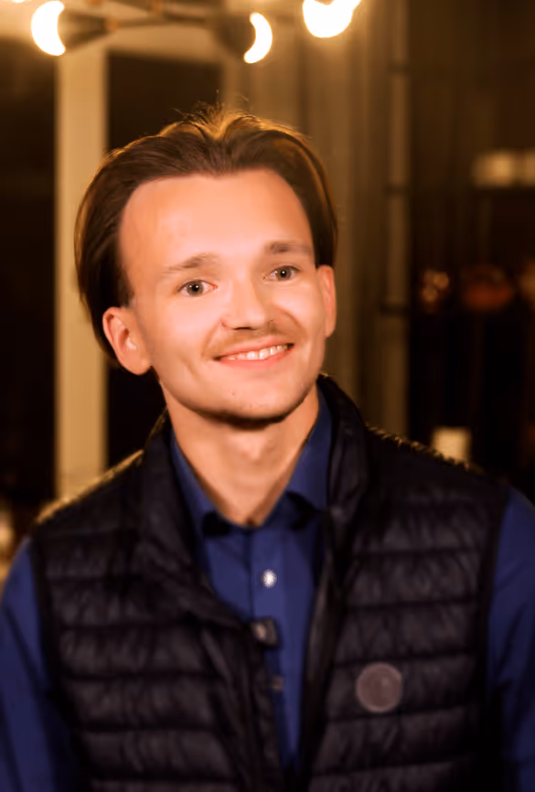 Junger Mann mit kurzer brauner Frisur, blaues Hemd und schwarzer gesteppter Weste lächelt in einem warm beleuchteten Innenraum.