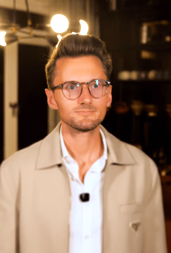 Junger Mann mit Brille und kurzem braunem Haar, trägt beige Jacke und weißes Hemd, steht vor unscharfem Hintergrund mit warmem Licht.