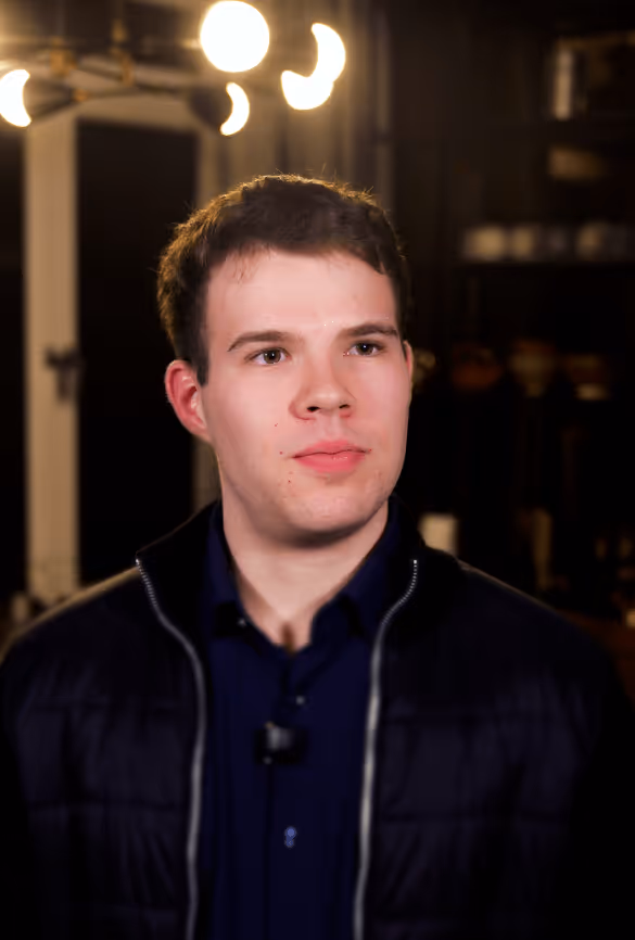 Ein junger Mann mit kurzen braunen Haaren trägt eine dunkle Jacke und ein blaues Hemd, im Hintergrund unscharfe Deckenleuchten.